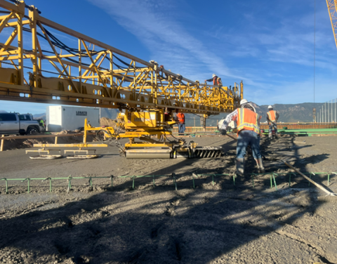 Construction workers using a large yellow machine to lay concrete on a road or parking lot. The machine has a long boom that extends over the workers, and they are using tools to smooth out the concrete. There are also some metal reinforcement bars visible in the concrete. The background shows a clear blue sky and mountains.