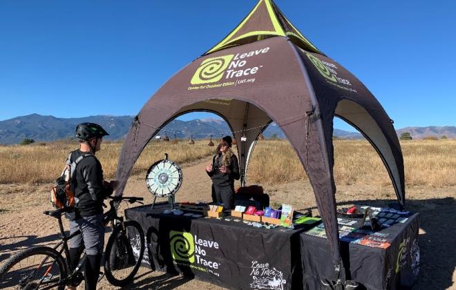 a person on a bike stops and talks to a Leave No Trace representative at a booth in an open space.