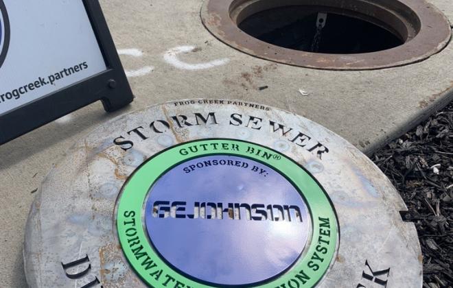 Lid to new storm drain where gutter bin is being installed. The words "storm sewer drains into creek. Gutter bin stormwater filtration system  sponsored by GE Johnson" is engraved on the lid.